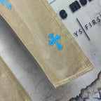 Beige pants leg with blue cross-heart patch near hem, straight cut