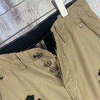 Tan pants with button closure and black cross patch embroidery, pocket close-up.