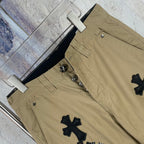 Tan cotton pants with button fly and black cross patches, close-up front.