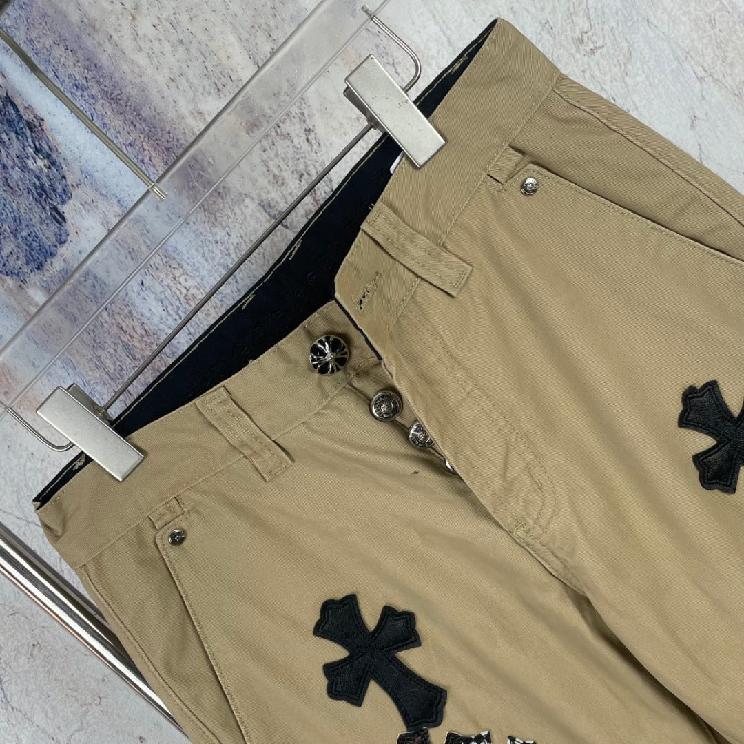 Tan cotton pants with button fly and black cross patches, close-up front.