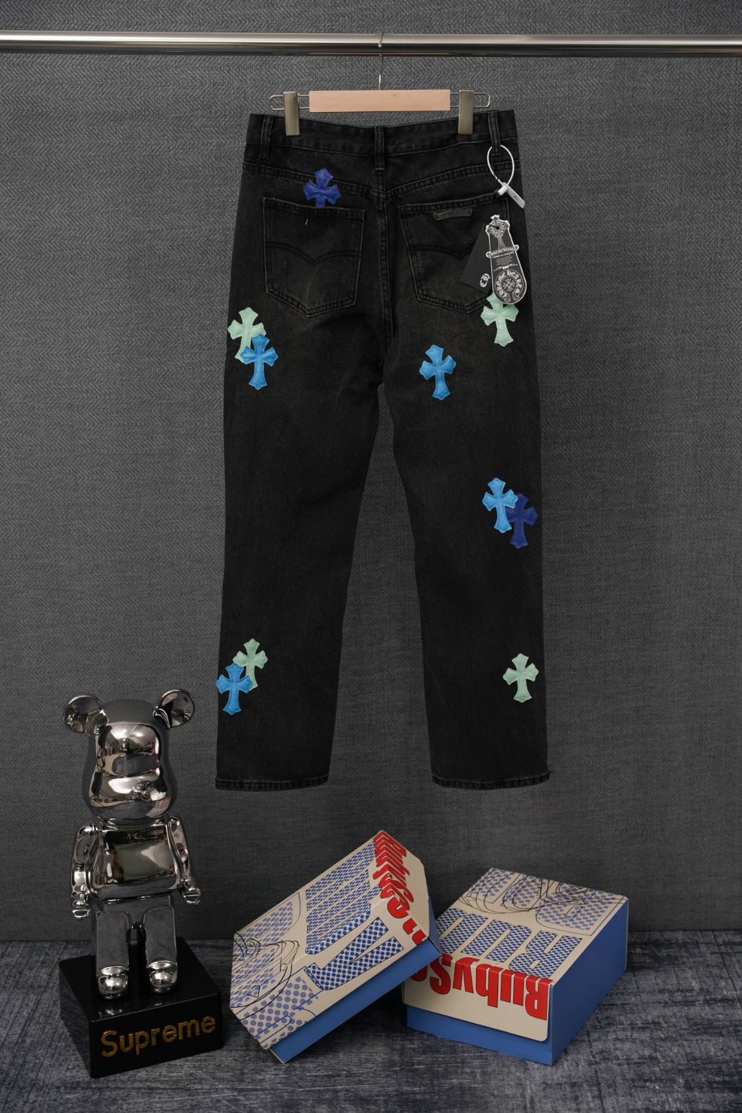 Black denim pants with colorful cross patches, back view, on hanger