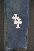 Blue denim pants leg with a single white cross patch near the hem.