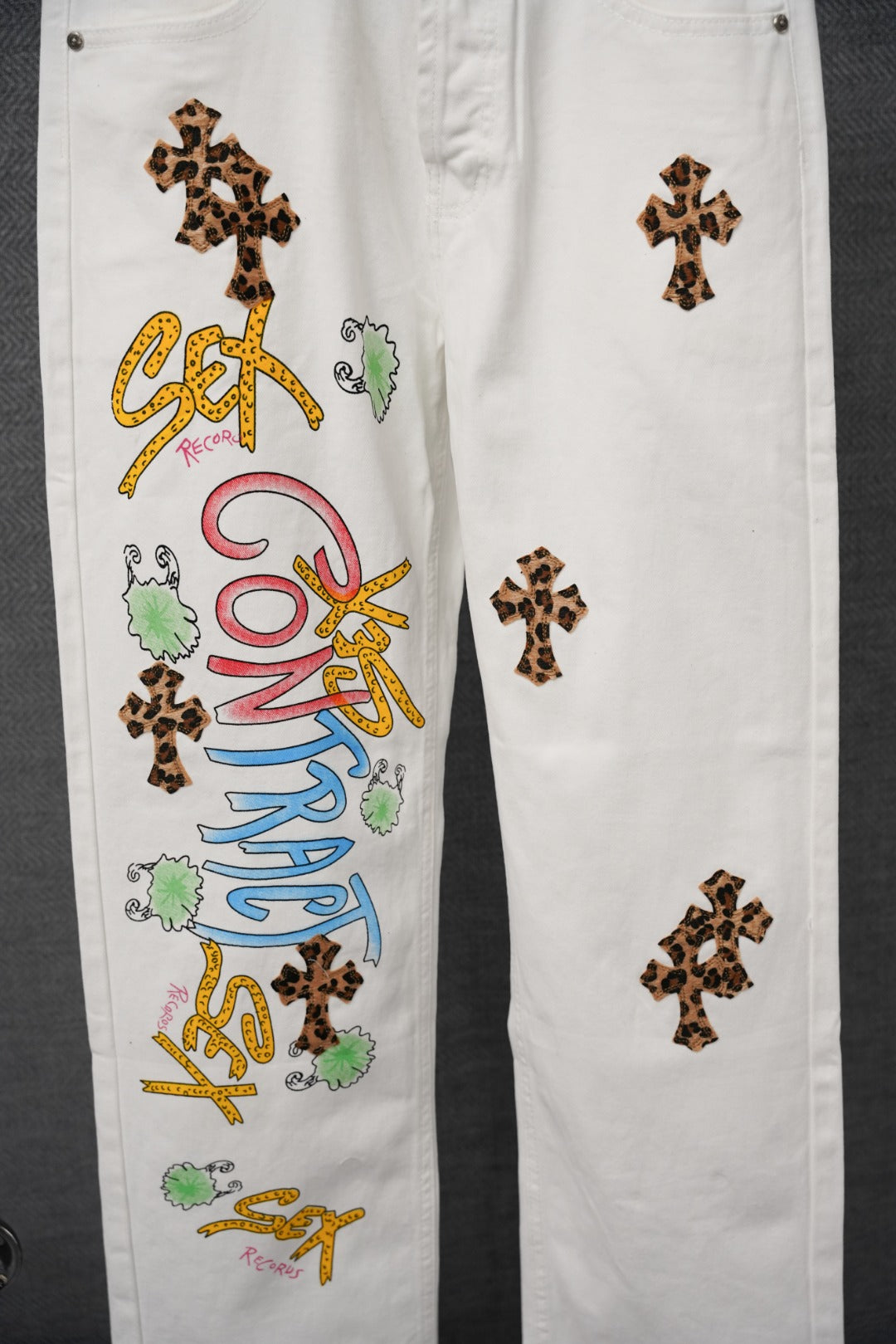 White denim pants with leopard-print cross patches and colorful vertical text graphic