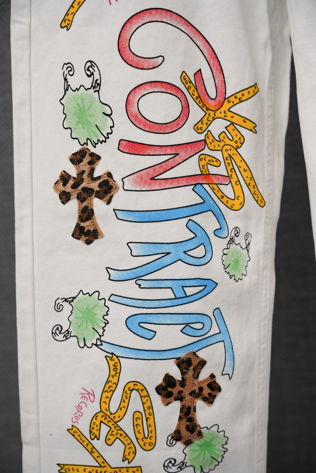 White denim pants with multicolor graphic lettering and leopard-print cross patches