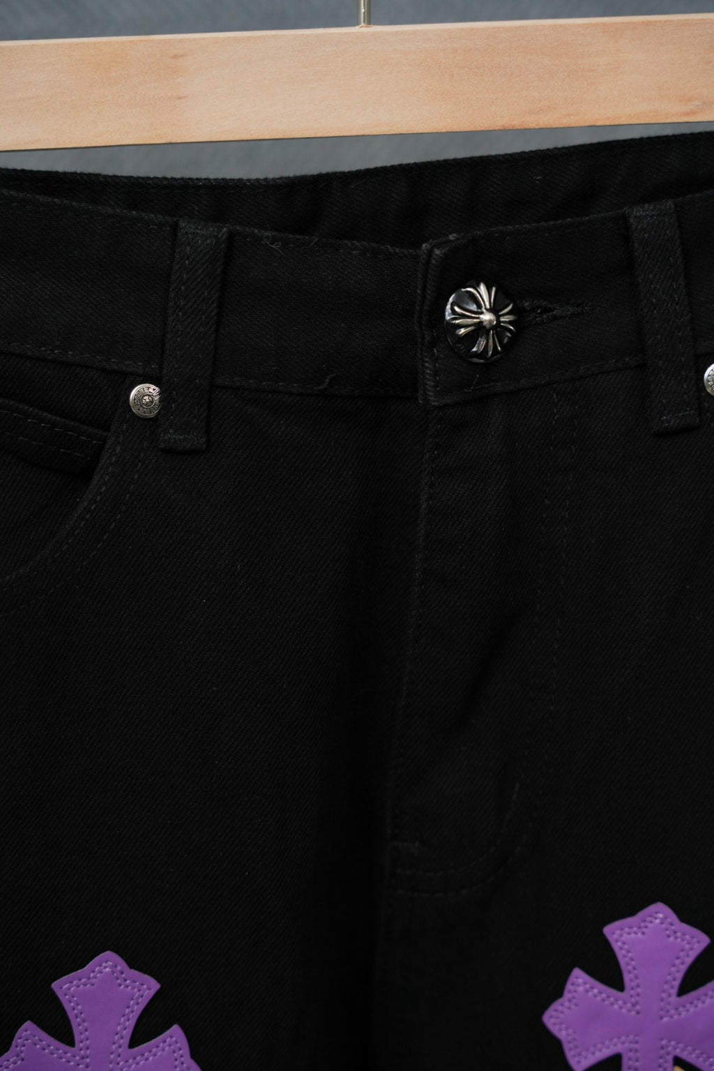 Black pants close-up: button fly, curved pocket seam, purple cross patch at hem.