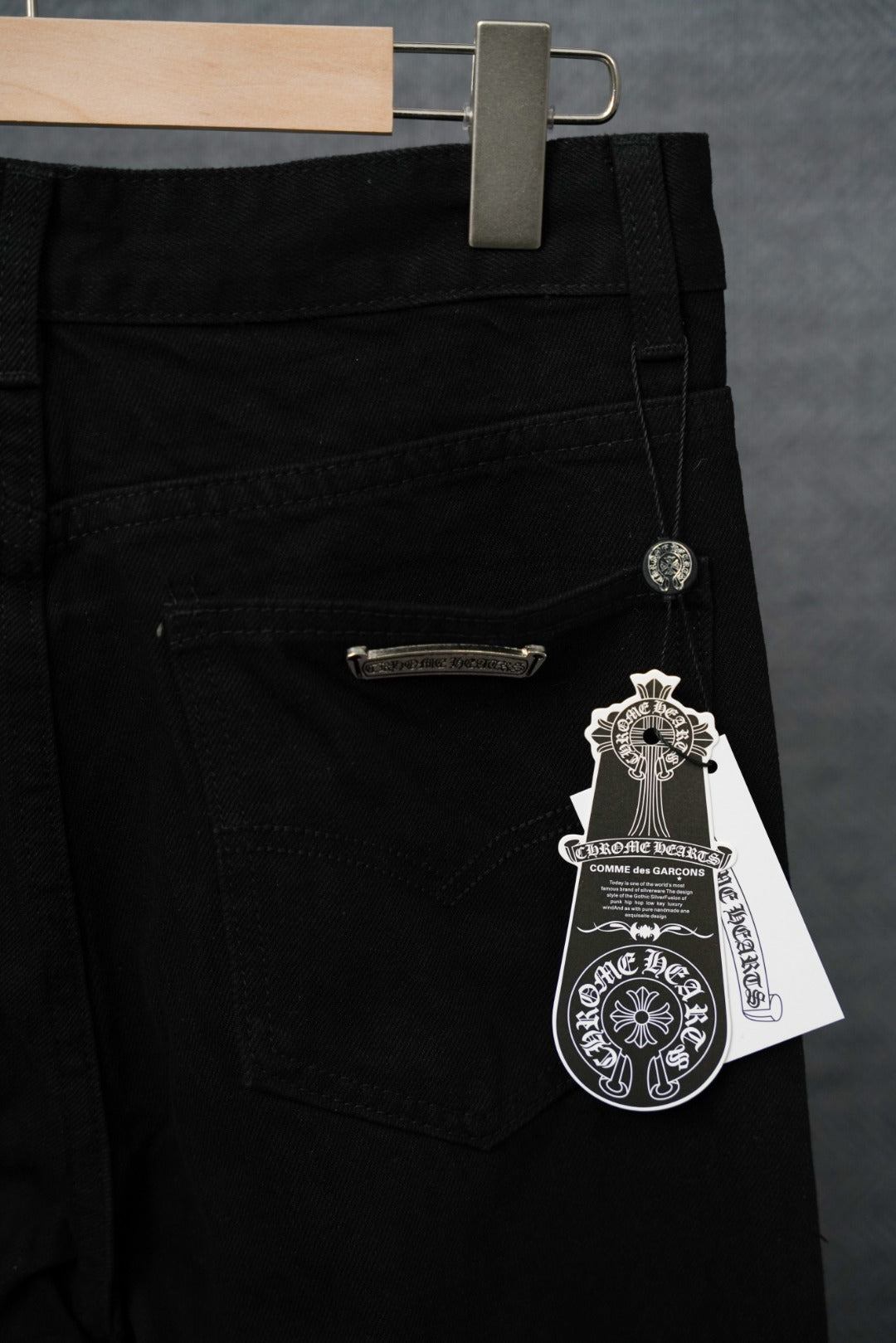 Black pants close-up: front pocket, zipper fly, silver logo charm and hanging tag.