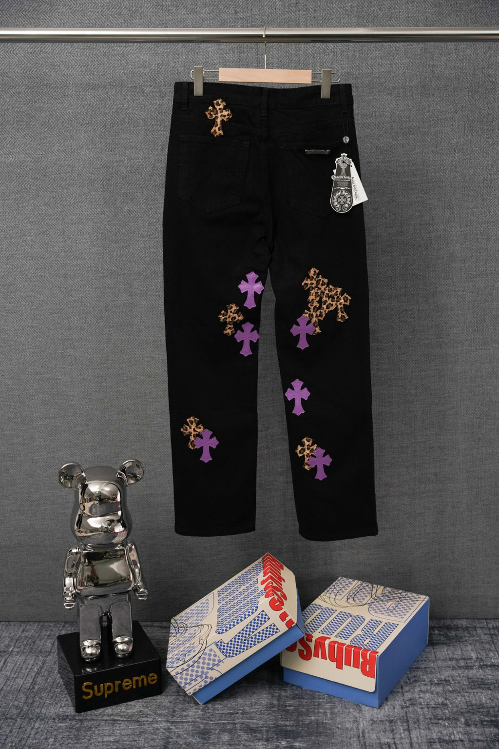 Black pants with scattered purple and gold cross patches, side view on hanger.