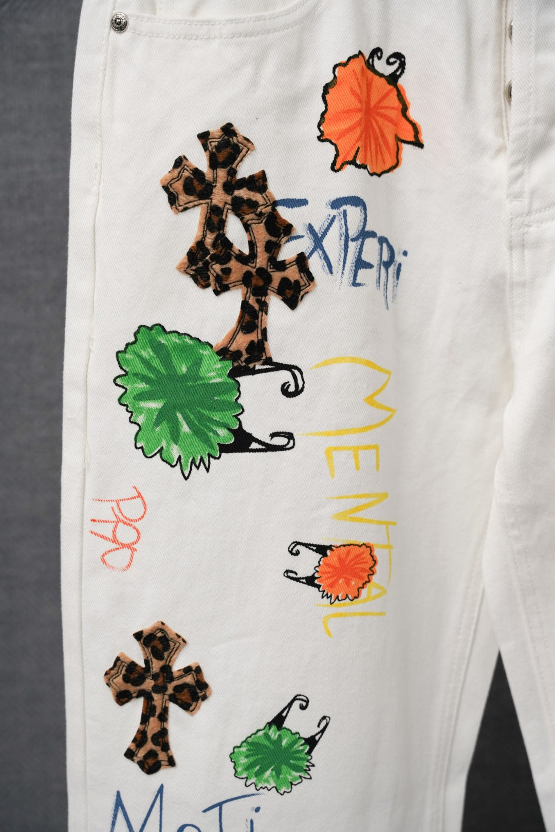 White graphic pants close-up showing leopard cross, orange dress, and green patches