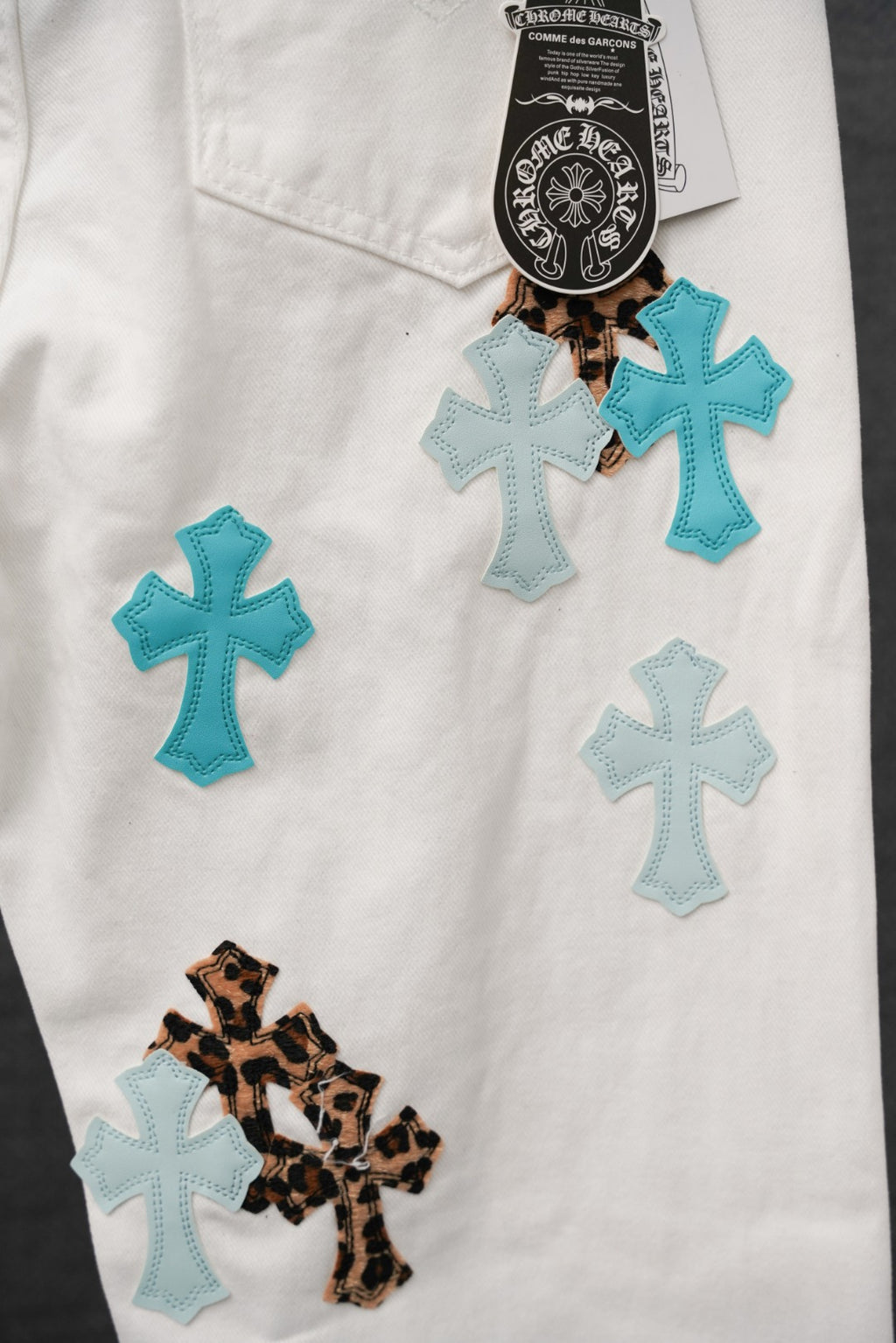 White pants with several teal crosses, one leopard cross patch, and brand tag