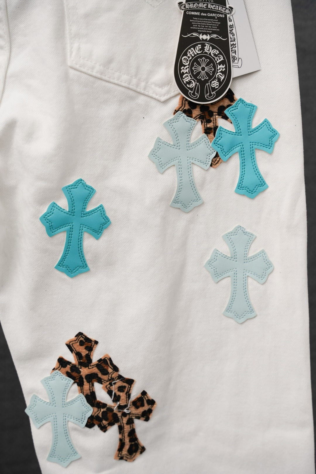 White pants with several teal crosses, one leopard cross patch, and brand tag