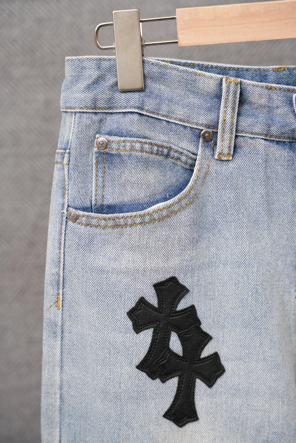 Light-wash denim pants front with button closure and a black cross patch