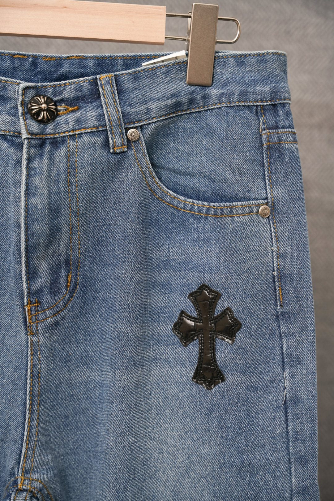 Light blue denim pants with button fly and black cross patch on lower front.