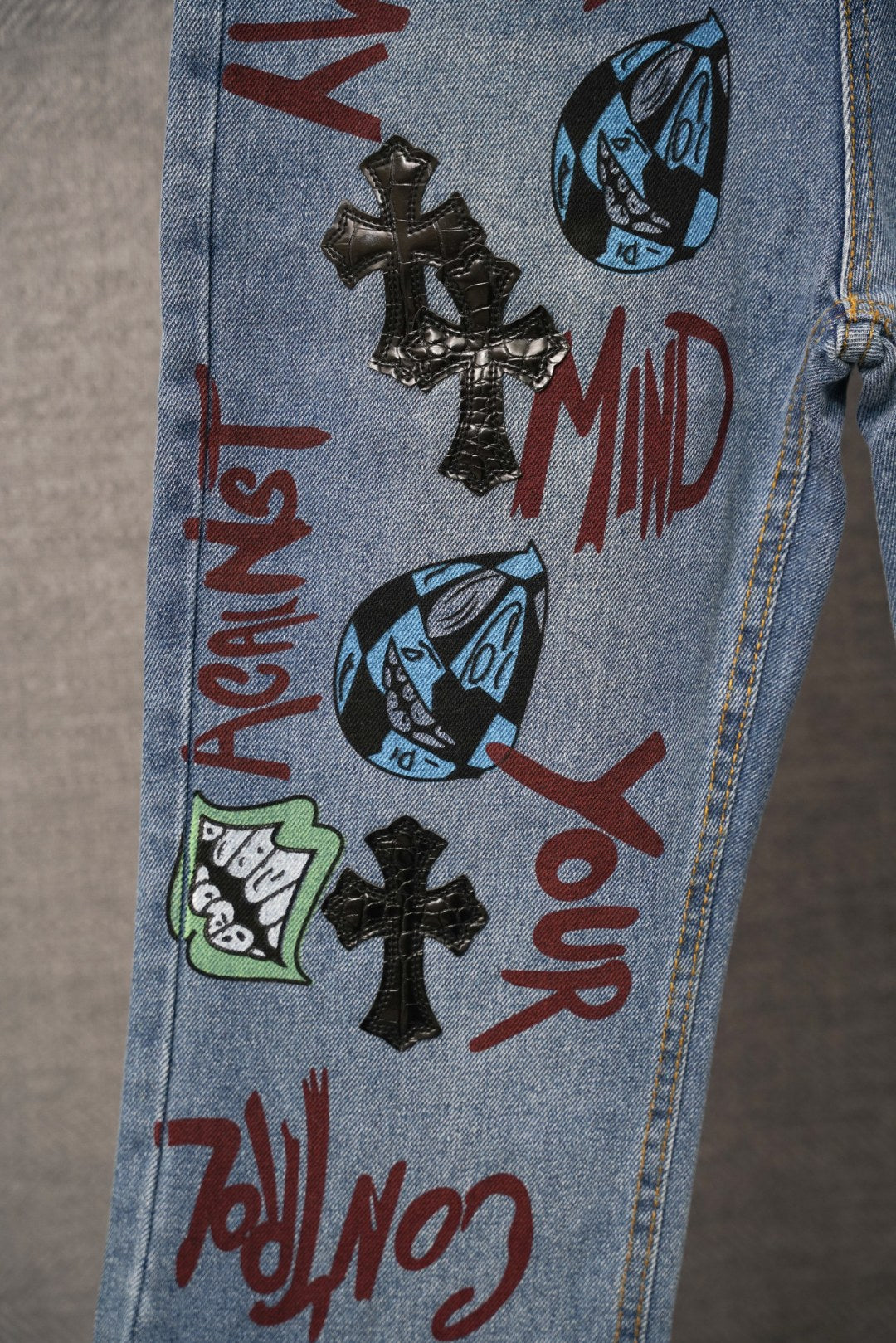 Light blue denim pants close-up of leg with graffiti text, faces, and black cross patch