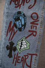 Light blue denim leg with vertical 'OVER' graffiti, cartoon faces, and black cross patch