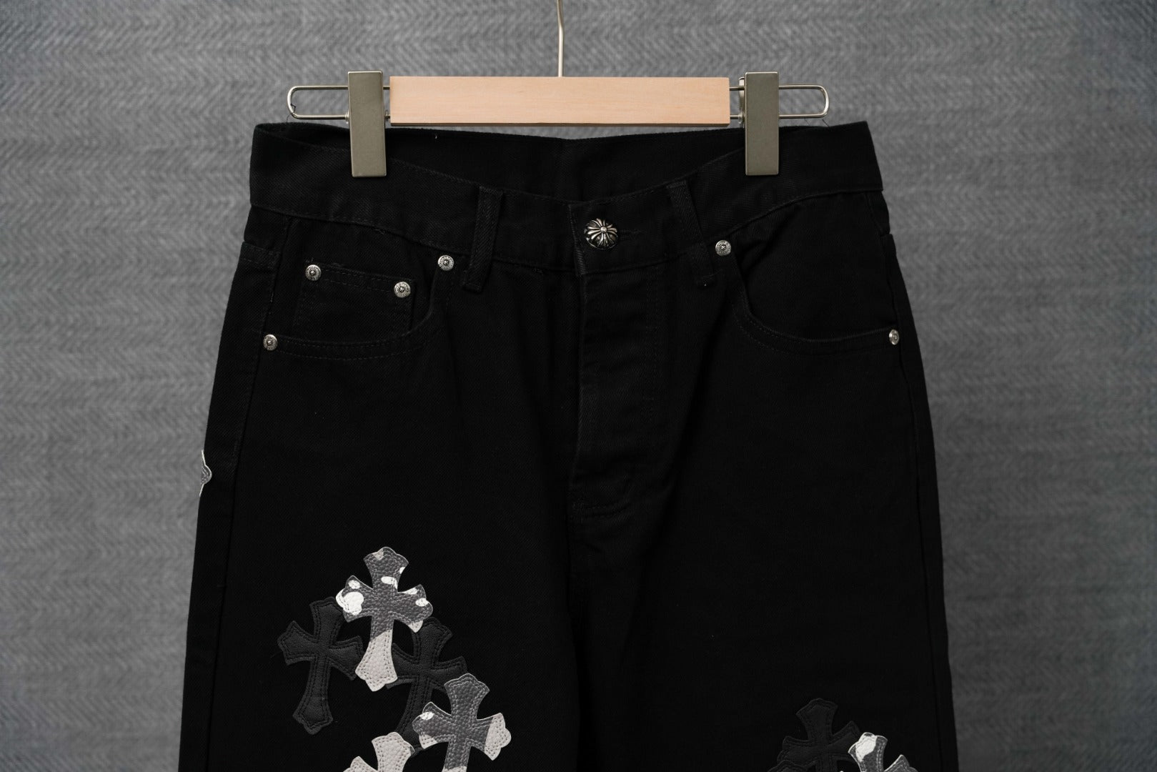 Black jean-style pants on hanger with button fly and silver cross appliqué on leg