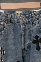 Light-wash denim jeans with front pleats and black cross appliqué near hem