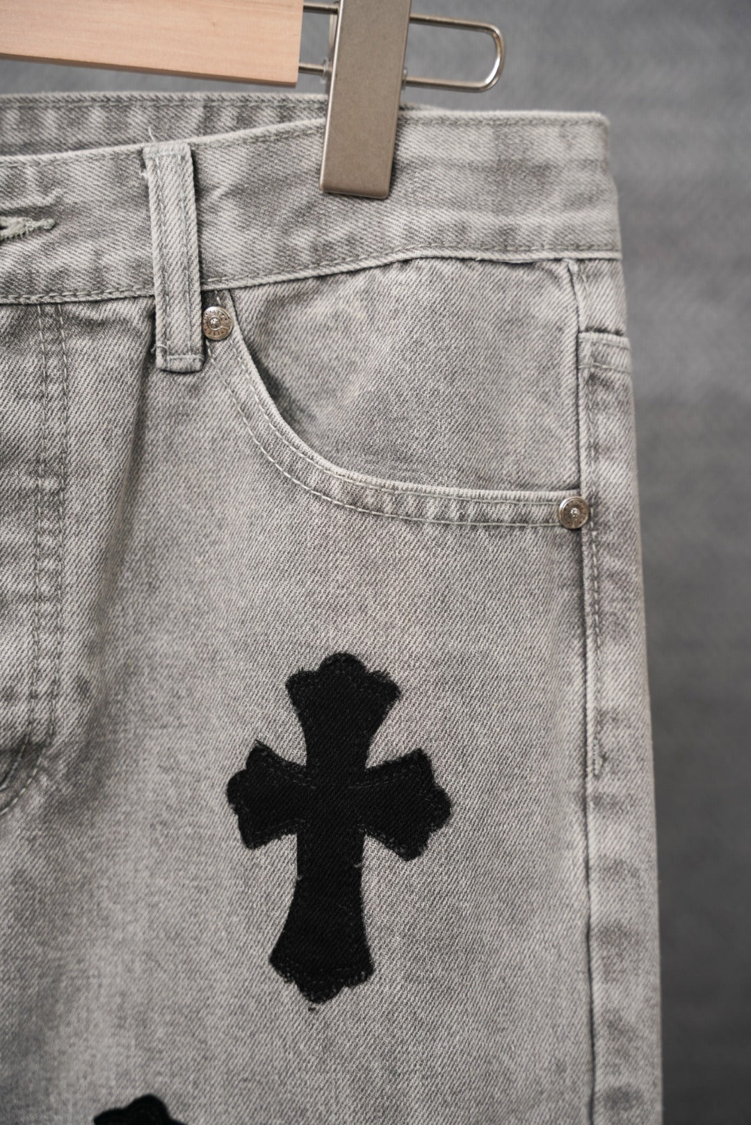 Light gray denim pants pocket and belt-loop area with black cross patch.