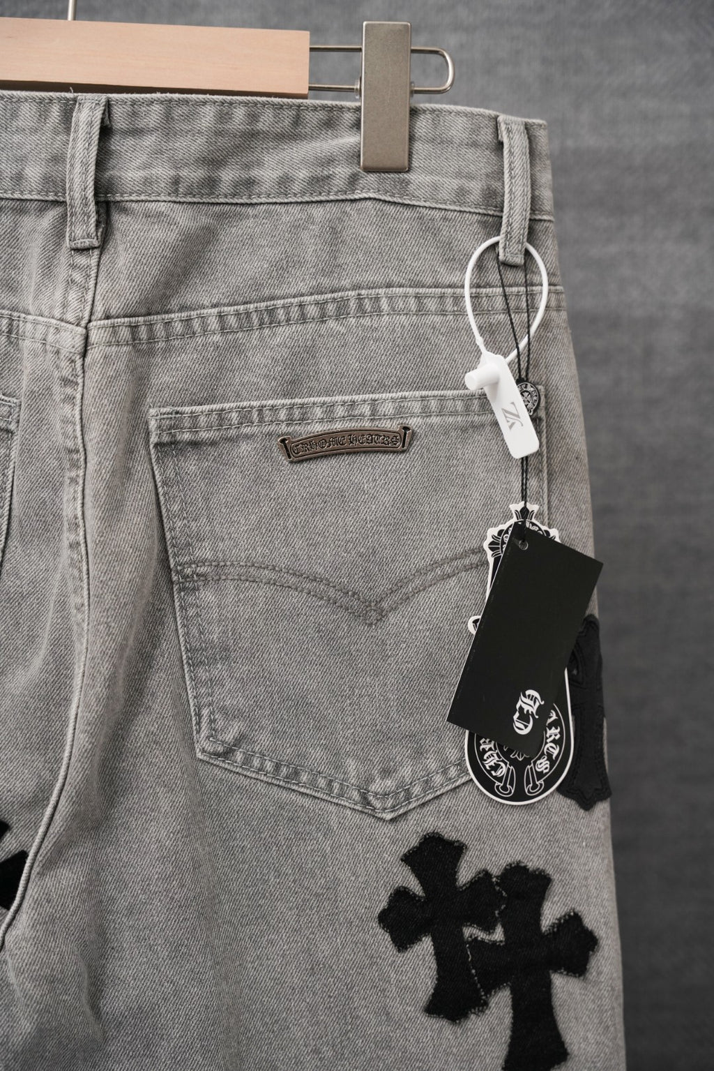Light gray denim pants with back pockets and black cross patch; hang tag visible.