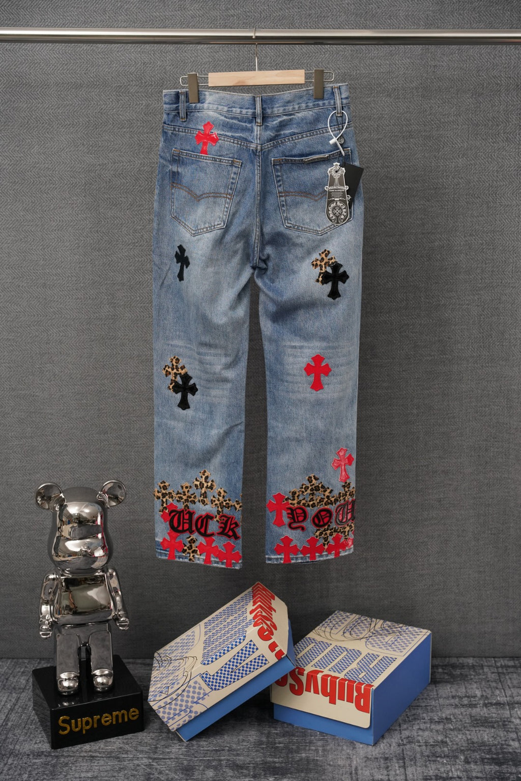 Light blue denim pants with cross patches and side keychain detail, rear view on hanger