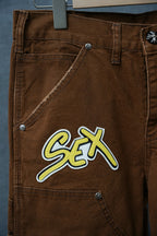 Brown cargo pants with neon “Sex” patch on thigh and metal buttons