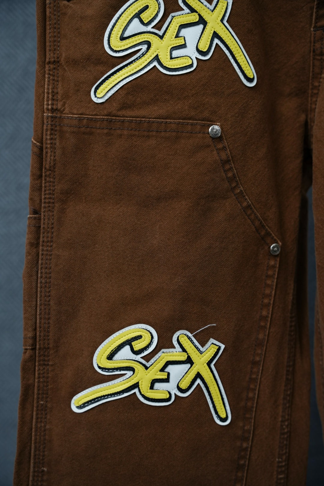 Brown cargo pants pocket with neon “Sex” patch and metal button