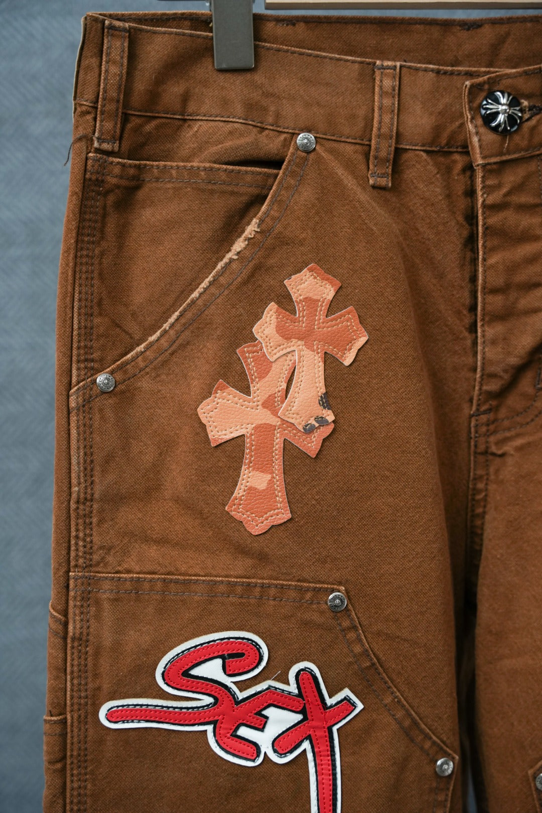Brown cargo pants close-up of side pocket with cross applique and “Sex” logo