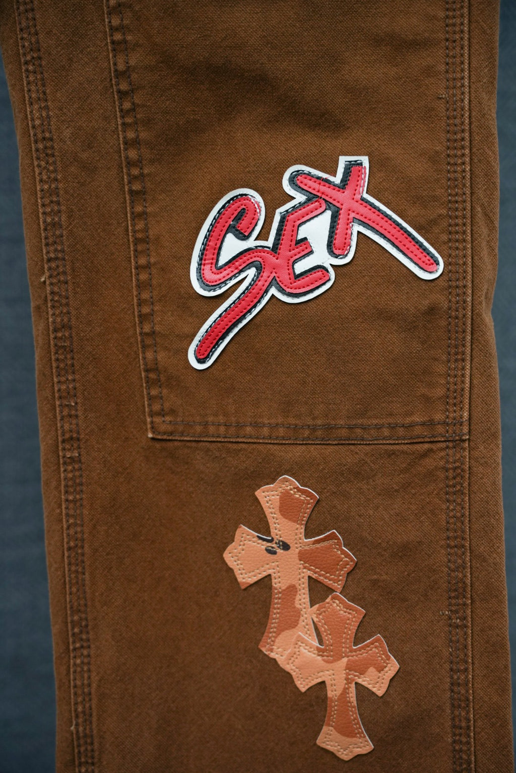 Brown cargo pants close-up of pocket graphic with “Sex” text and cross applique below