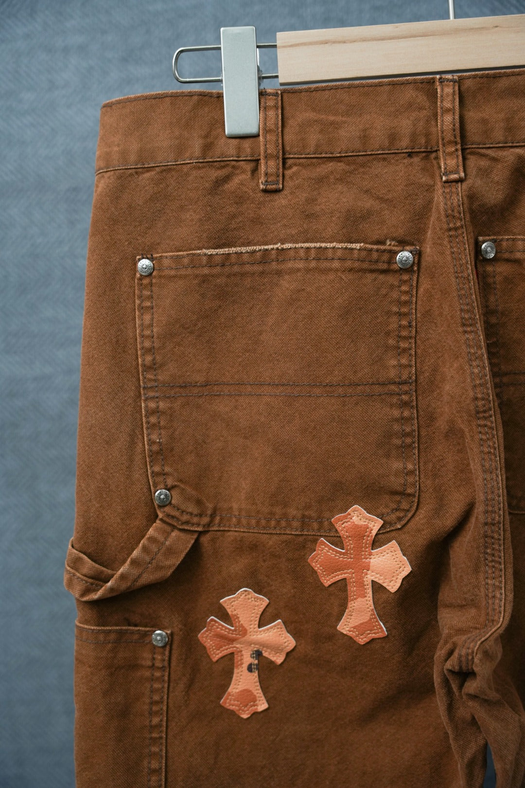 Brown chrome pants with buttoned waistband and orange cross patch on pocket