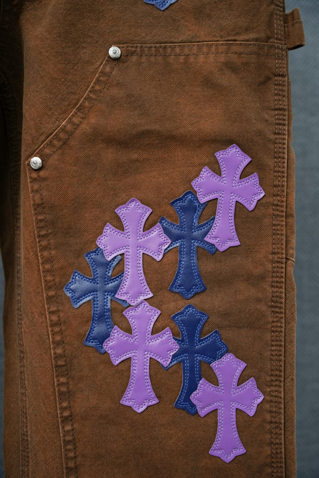 Brown denim pants with row of blue and purple cross patches on leg