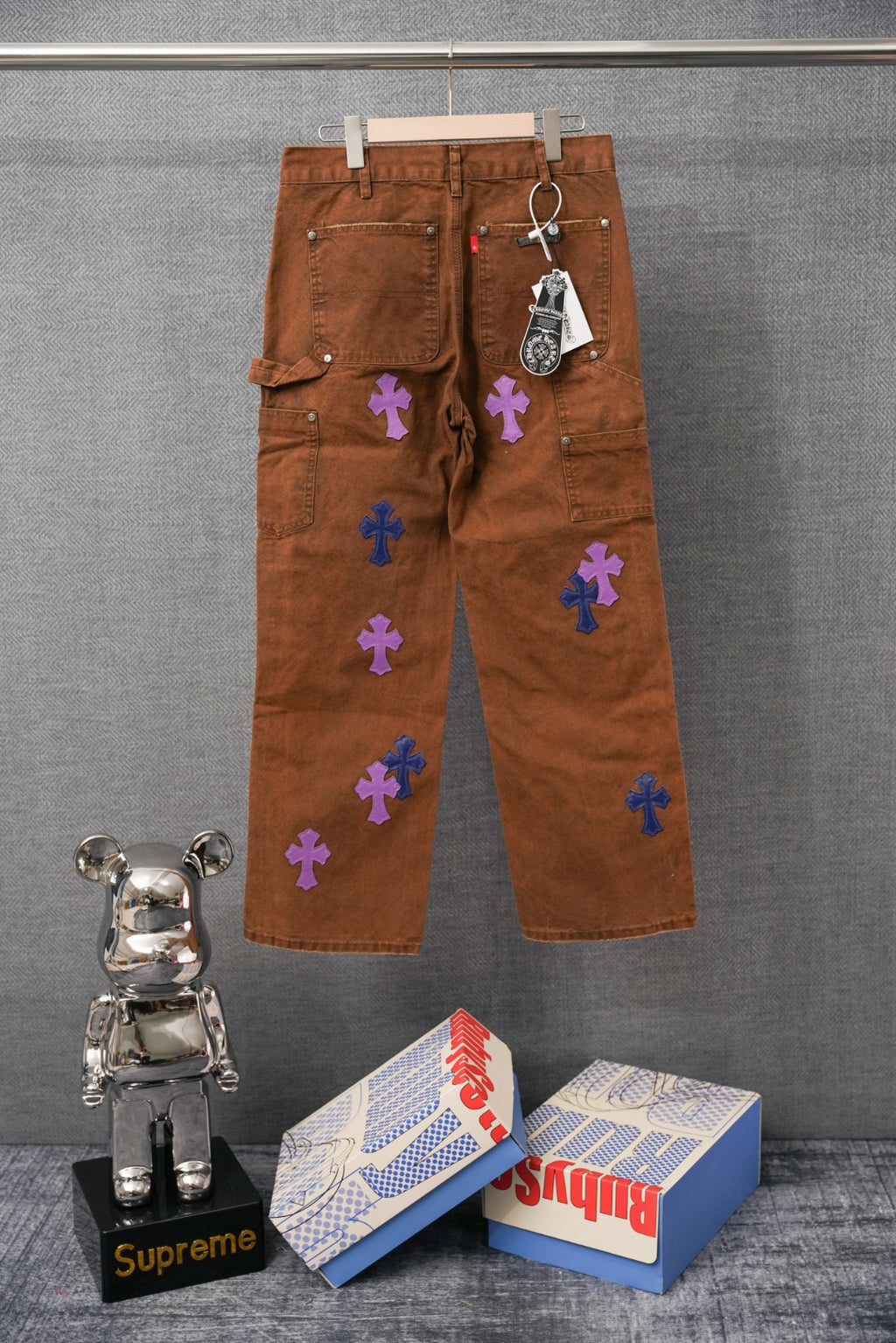 Brown denim pants with cross patches in blue and purple, side view on hanger