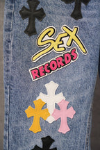 Blue denim pants with 'Sex Records' patch and black cross graphics, close-up.