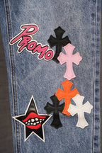 Blue denim pants with 'Promo' patch and pink and black cross graphics, close-up.