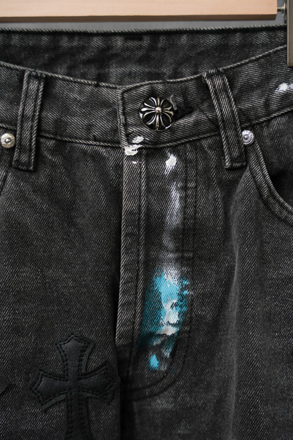 Black denim pants waistband with engraved button and cross-themed patch detail